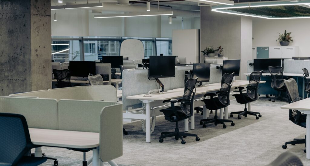 The image depicts a modern, open-plan office with a clean and organized layout. The workspace features multiple desks equipped with computers and ergonomic chairs. The desks are separated by low dividers, creating individual workstations while maintaining an open and collaborative environment. Overhead lighting and large windows provide ample illumination, enhancing the bright and airy atmosphere of the office. The decor includes minimalistic elements with a few potted plants adding a touch of greenery. The overall design emphasizes functionality and comfort, making it a conducive environment